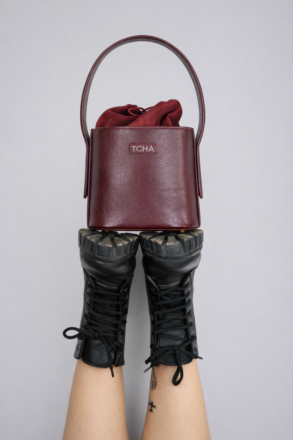 Burgundy handbag with 'TCHA' branding held by a person wearing black boots on a gray background